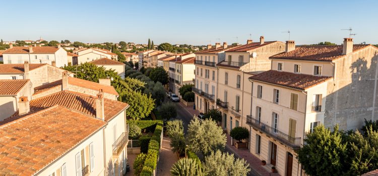 Vue immobilière réaliste de Béziers avec maisons résidentielles, toits en terre cuite, immeubles du centre ancien, rues ensoleillées et végétation méditerranéenne.